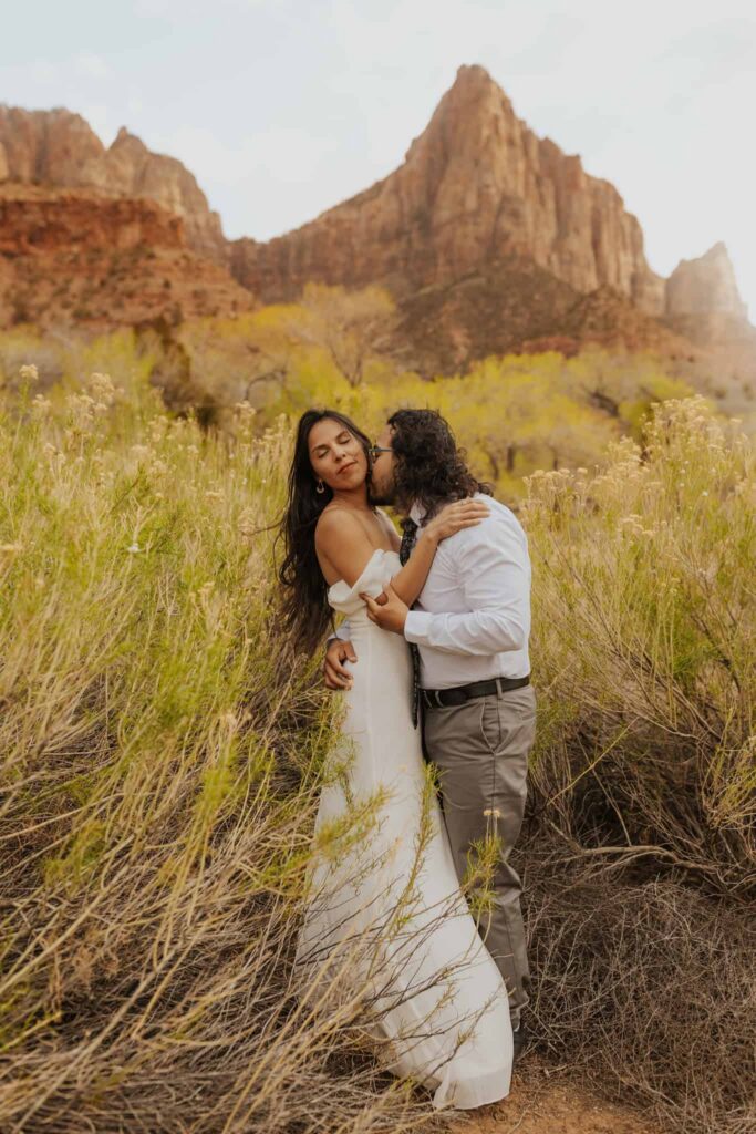A groom kissing his bride on the cheek next to a spring time flowers in Zion